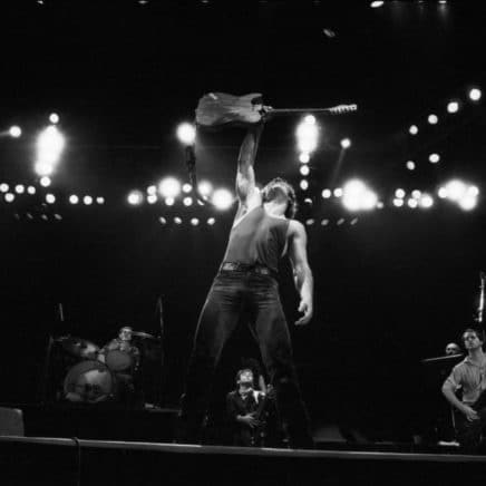 Bruce Springsteen holds his electric guitar triumphantly over his head with one hand. He is on stage at a concert with lights and other musicians behind him.