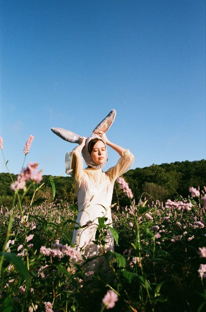 Musician Charlotte Bumgarner stands in a field of flowers. She is adjusting bunny ears that are on her head.