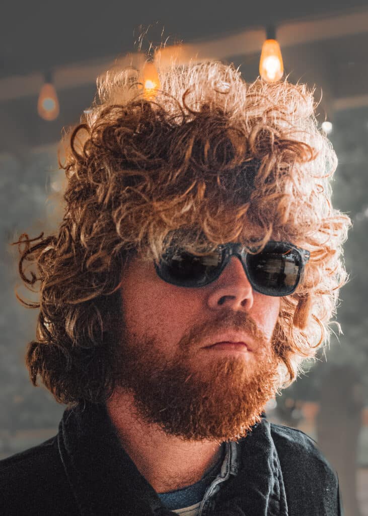 Musician JW Francis looks sternly at the camera. His hair is significantly curly and he wears wayfarer-style sunglasses.