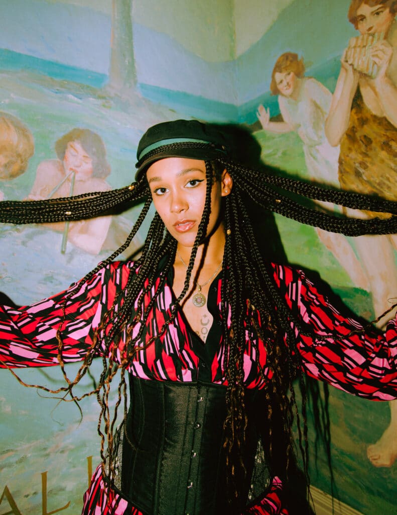 Musician LEX stands in front of an idyllic, painted mural. She holds holds long braids out horizontally and looks directly at the camera.